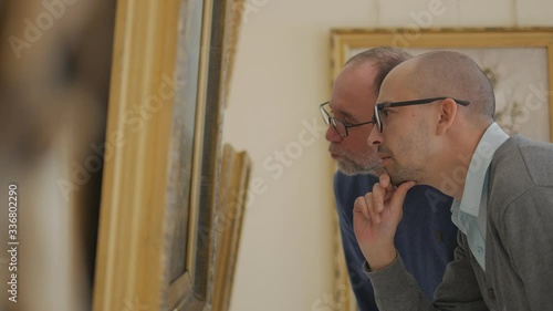 Two men in an art gallery look at a painting and talk about art
