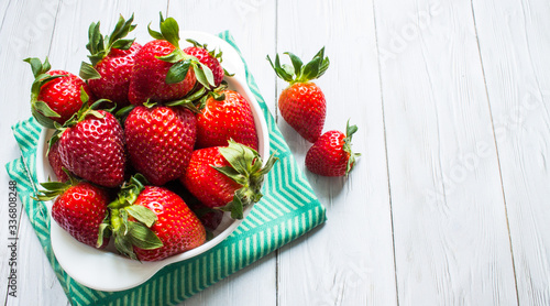 Bright red strawberry berries in a white Cup close up with green textiles on a white wooden background with space top view