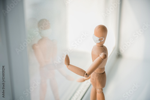 Wooden mannequin in medical mask in front of the window. Concept of isolation and staing home during the quarantine.