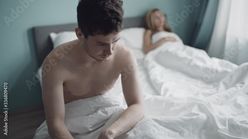 Problem in relationship. Unhappy couple. Worried thoughtful man sitting apart on bed, feeling sad, wife in background, thinking of breaking up, divorce. Impotence, quarrel, family, sex coflict concept photo