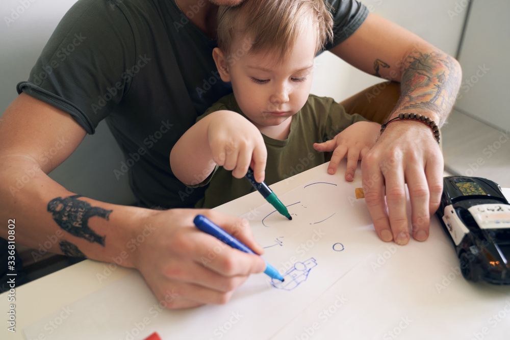 Young father helps son at age of two draw, teaches alphabet. A small boy is sitting on his dads lap near white wall in room at table, on piece of paper writing letters and pictures with felt-tip pen. Young father helps son at age of two draw, teaches alphabet. A small boy is sitting on his dads lap near white wall in room at table, on piece of paper writing letters and pictures with felt-tip pen.