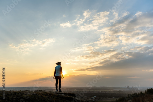 Silhouette of a woman hiker standing alone enjoying sunset outdoors. Female tourist on rural field in evening nature. Tourism, traveling and healthy lifestyle concept.