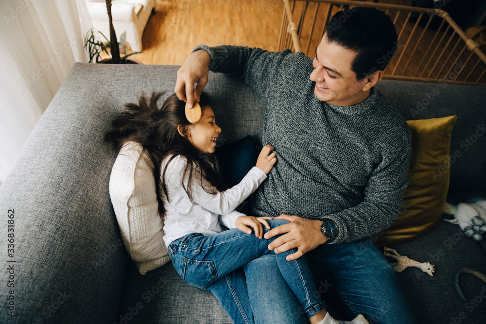 High angle view of father combing daughter's hair while sitting on sofa at home High angle view of father combing daughter's hair while sitting on sofa at home