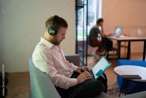 Caucasian man in electronic equipment listening medial playlist from music platform and typing text during web chatting in social networks, skilled freelancer in headphones for noise cancellation