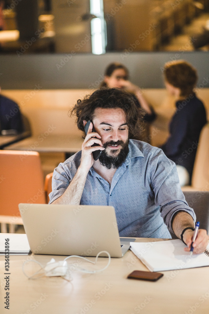 Businessman writing in diary while talking on smart phone in office Businessman writing in diary while talking on smart phone in office