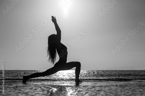 Silhouette of woman standing at yoga pose on the tropical beach during sunset. Girl practicing yoga near sea water. Black and white