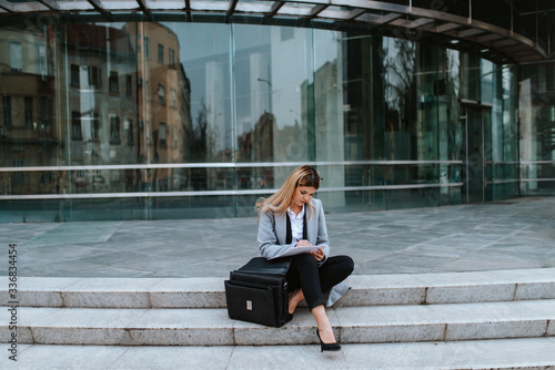 A young attractive businesswoman with a bag is sitting on the stairs in front of the company © Dragica