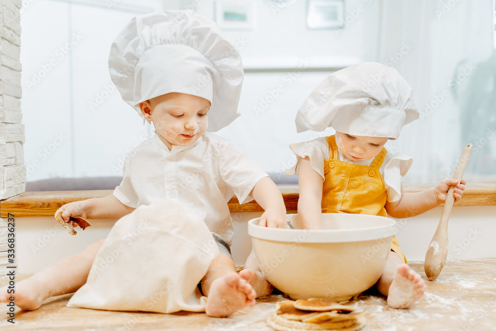 Charming two-year-old twins brother and sister in chef's hats are mixing pastry for pies while sitting on a table in the kitchen. Concept of children's hobbies and work for children at home Charming two-year-old twins brother and sister in chef's hats are mixing pastry for pies while sitting on a table in the kitchen. Concept of children's hobbies and work for children at home