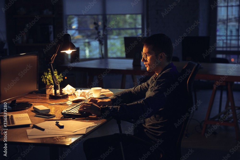 Married to his job. Man working in office alone during COVID-19 quarantine, staying to late night. Young businessman, manager doing tasks with smartphone, laptop, tablet in empty workspace.