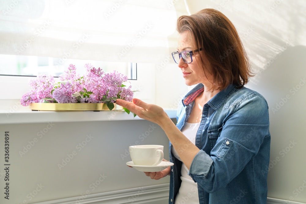Smiling female standing near window with cup of tea and bouquet of lilacs Smiling female standing near window with cup of tea and bouquet of lilacs