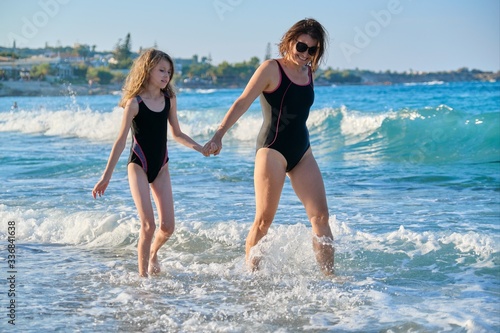 Mother and daughter child in swimsuits walking along seashore holding hands