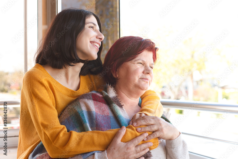 Happy senior mother and adult daughter looking at the view through the window.