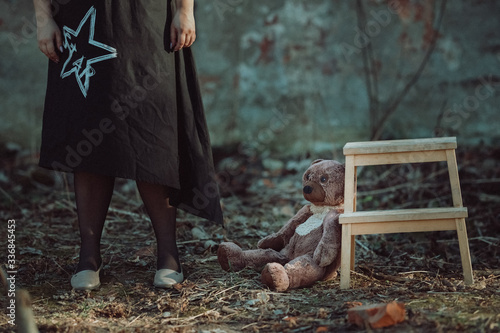 girl stands next to a teddy bear sitting on the ground near a wooden chair in the middle of the ruins of a brick building post apocalyptic concept © Mk16.15