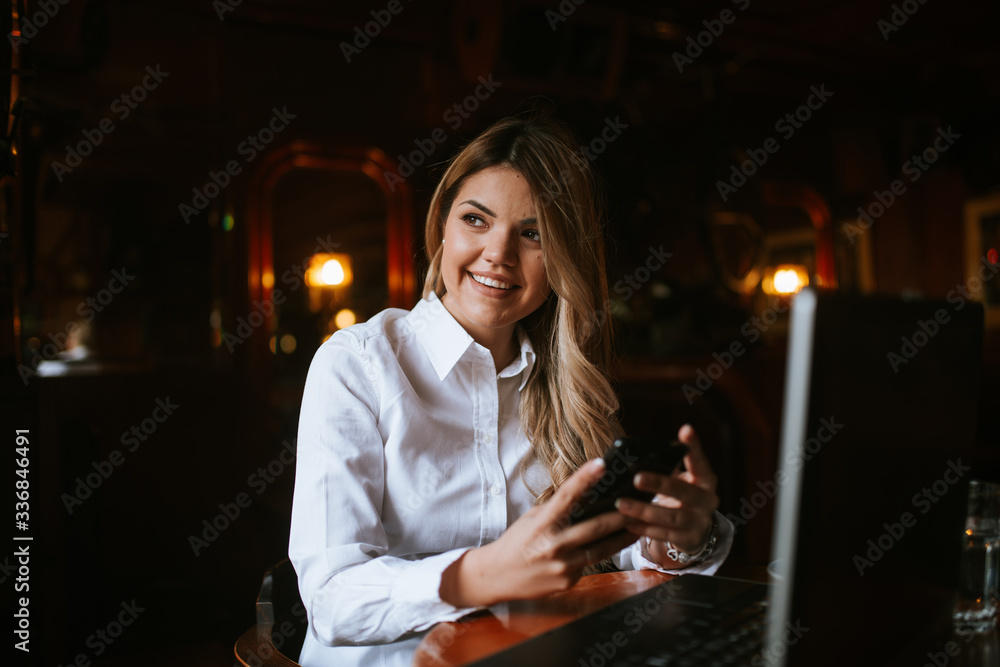 A young attractive caucasian  blond girl with a phone is sitting in a cafe in front of a laptop