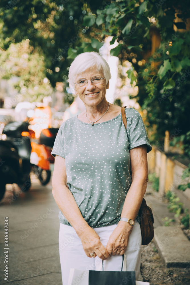 Portrait of smiling senior woman with bag standing on sidewalk in city Portrait of smiling senior woman with bag standing on sidewalk in city