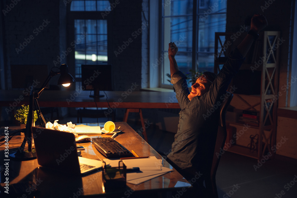 Done with deadline. Man working in office alone during coronavirus or COVID-19 quarantine, staying to late night. Young businessman, manager worked with smartphone, laptop, tablet in empty workspace. Done with deadline. Man working in office alone during coronavirus or COVID-19 quarantine, staying to late night. Young businessman, manager worked with smartphone, laptop, tablet in empty workspace.