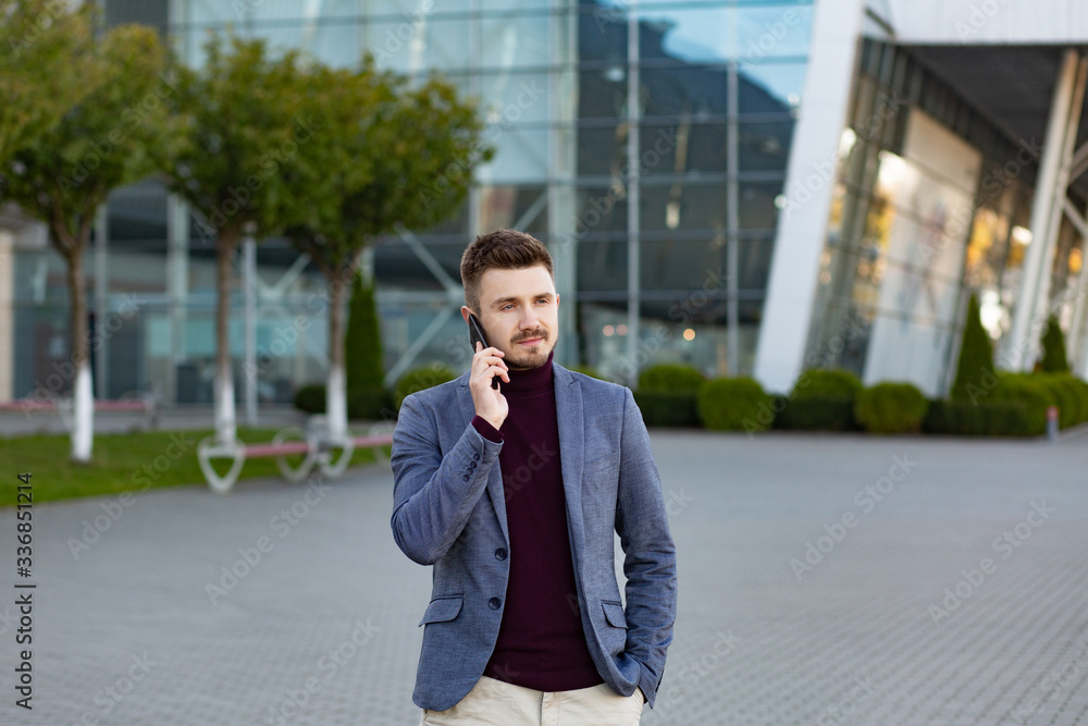 Portrait of handsome serious man in casual wear holding smartphone. Successful manager makes a call. Young businessman talking on the phone, standing on city street near business center. Portrait of handsome serious man in casual wear holding smartphone. Successful manager makes a call. Young businessman talking on the phone, standing on city street near business center.