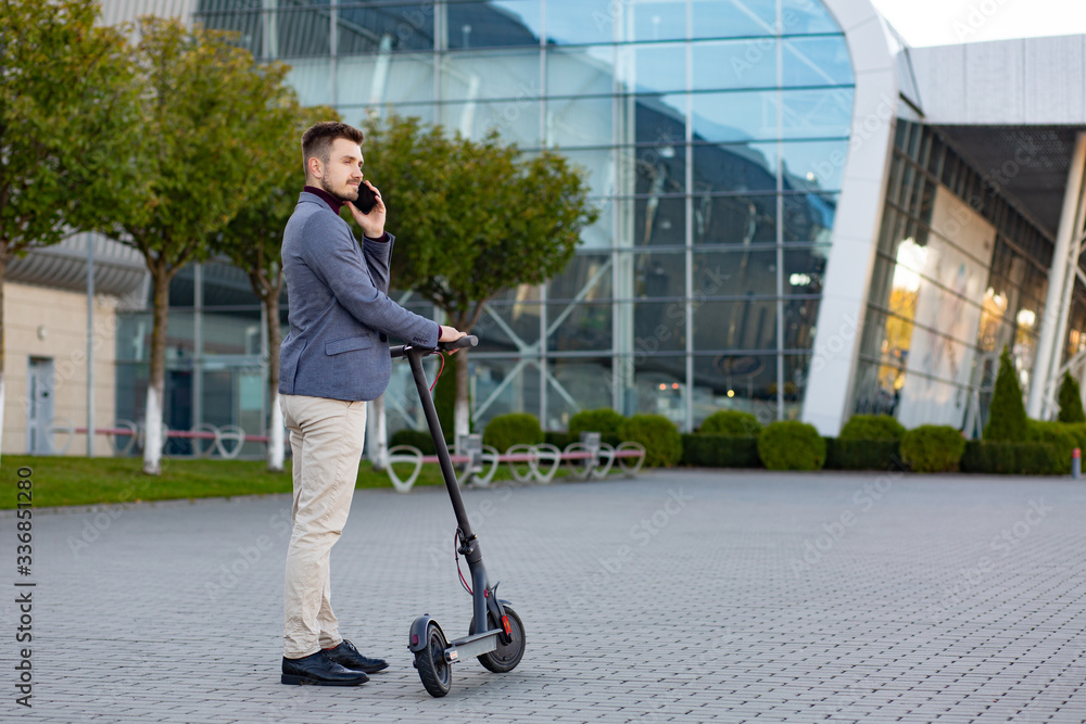 Young handsome man with e-scooter standing on sidewalk near the airport on the sunset and talking on the smartphone.Trendy urban transportation on modern electric scooter.Eco friendly mobility concept Young handsome man with e-scooter standing on sidewalk near the airport on the sunset and talking on the smartphone.Trendy urban transportation on modern electric scooter.Eco friendly mobility concept