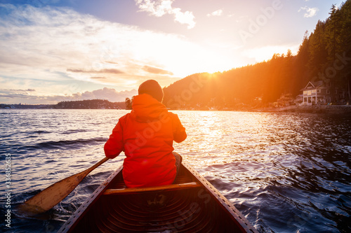 Adventurous Man on wooden canoe are paddling in water during a vibrant sunny sunset. Taken in Indian Arm, near Deep Cove, North Vancouver, British Columbia, Canada. Concept: Adventure, Sport, Explore