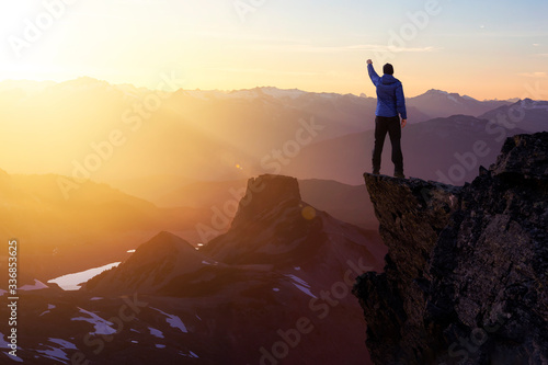 Composite. Adventurous Man Hiker With Hands Up on top of a Steep Rocky Cliff. Sunset or Sunrise. Landscape Taken from British Columbia, Canada. Concept: Adventure, Explore, Hike, Lifestyle