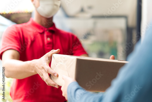 Asian postman, deliveryman wearing mask carry small box deliver to customer in front of door at home. Man wearing mask prevent covid19, corana virus affection outbreak. Social distancing work concept. © Kawee