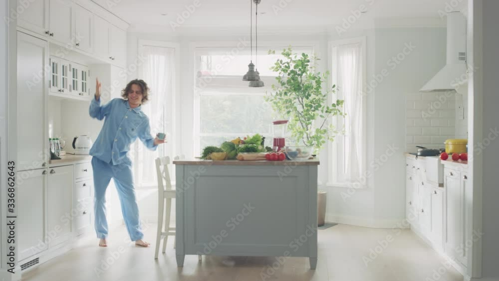 Happy Young Man with Long Hair Dancing at Home while Wearing Blue Pajamas. He is Listening to Music on a Mobile. Energetic Man Using Smartphone in a Kitchen with Healthy Vegetables. Happy Young Man with Long Hair Dancing at Home while Wearing Blue Pajamas. He is Listening to Music on a Mobile. Energetic Man Using Smartphone in a Kitchen with Healthy Vegetables.