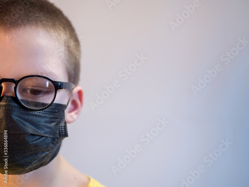 cute blonde boy in black medical mask and glasses is quarantined at home. child coughs heavily and wears mask. concept of fight against the coronavirus epidemic and proper prevention of infections