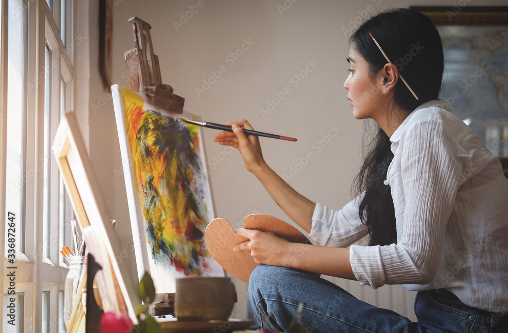 Cropped shot view of Woman painter sitting on the floor in front of the canvas and drawing. Artist studio interior. Drawing supplies, oil paints, artist brushes, canvas, frame. - obrazy, fototapety, plakaty 