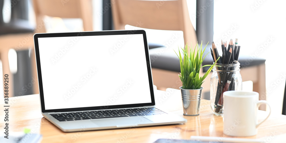 Fototapeta premium Blank screen computer laptop putting on wooden working desk and surrounded by potted plant, pencil holder and ceramic coffee cup. Orderly workspace and space for advertisement concept.