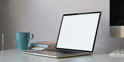 Photo of white blank screen computer laptop putting on white working desk surrounded by computer monitor, coffee cup and stack of books over white wall as background.