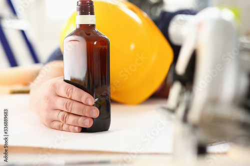 Arm of drunken worker in yellow helmet hold liquor bottle sleeping at table closeup. Manual job workplace DIY inspiration fix shop hard hat industrial education profession career