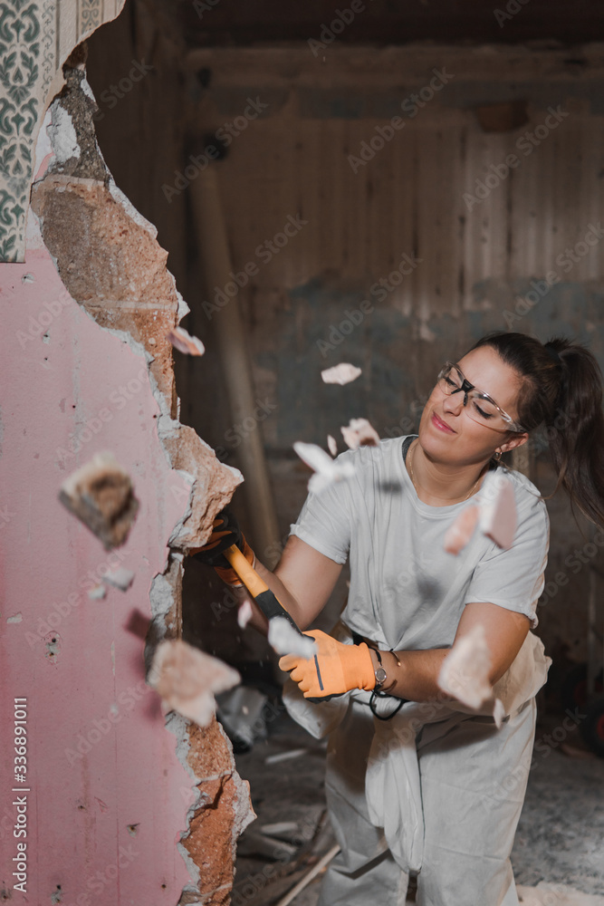 Young very strong and tough woman in white working outfit clothes hitting hard and breaking a wall with a large heavy hammer  during manly renovation work and pieces of the wall flying away