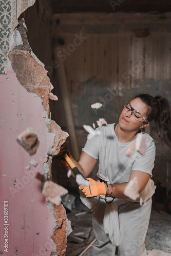 Fototapeta Naklejka Na Ścianę i Meble -  Young very strong and tough woman in white working outfit clothes hitting hard and breaking a wall with a large heavy hammer  during manly renovation work and pieces of the wall flying away