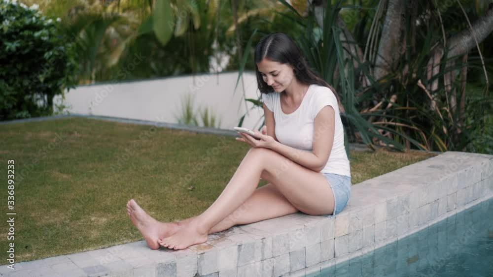 Young woman sits on edge of pool and uses phone for work, greneery on background Young woman sits on edge of pool and uses phone for work, greneery on background