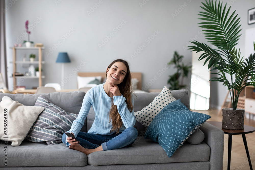 Smiling woman using mobile on the sofa at home. Online chat. - obrazy, fototapety, plakaty 