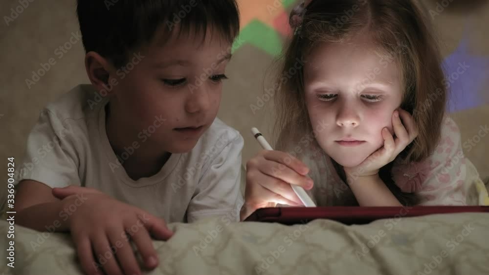 Little children, brother and sister are lying on the bed and playing on the tablet before going to bed. Little children, brother and sister are lying on the bed and playing on the tablet before going to bed.