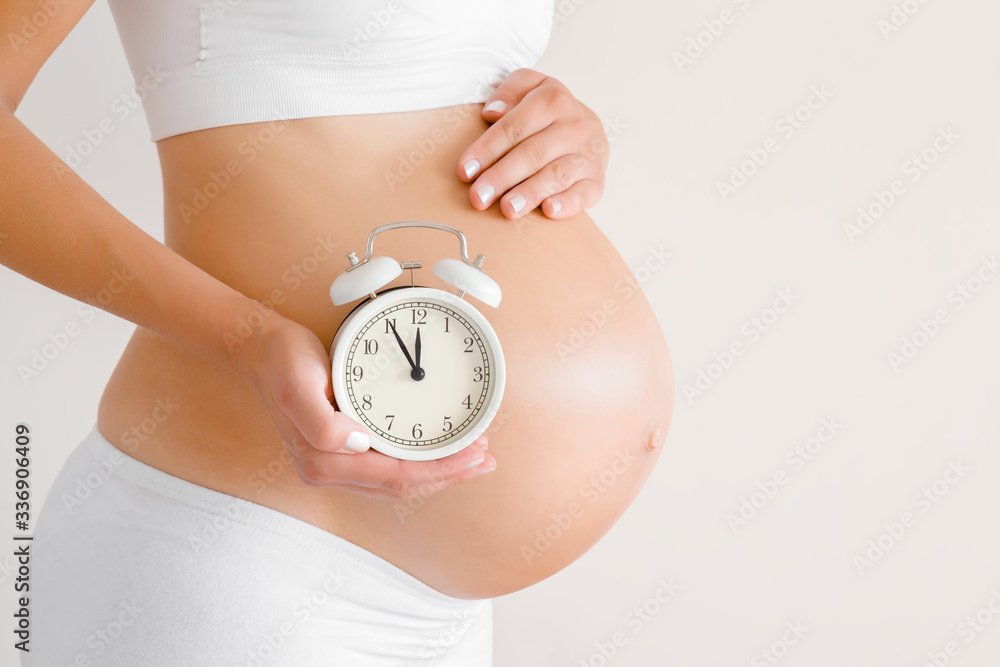Young woman standing and touching naked big belly. Hand holding white alarm  clock. Baby expectation. Emotional loving pregnancy time - 37 weeks.  Childbirth time. Closeup. Isolated on gray background. Stock Photo