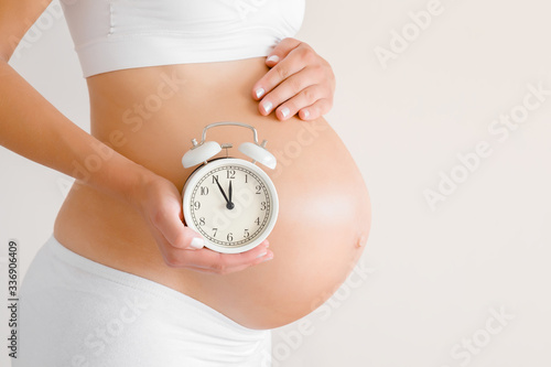 Young woman standing and touching naked big belly. Hand holding white alarm clock. Baby expectation. Emotional loving pregnancy time - 37 weeks. Childbirth time. Closeup. Isolated on gray background.