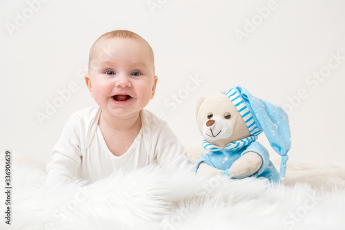 Happy just awake baby girl in white clothes with sleepy teddy bear in blue pajama and hat sitting on fluffy fur blanket. Kids best friend for sweet dreams. Front view. Closeup.