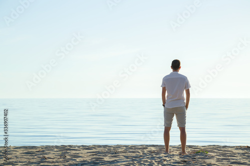 Fototapeta Naklejka Na Ścianę i Meble -  Young man in white clothes standing alone on sand and staring at calm sea and light blue sky. Fresh air. Empty place for emotional, sentimental, inspirational text, quote or sayings. Back view.