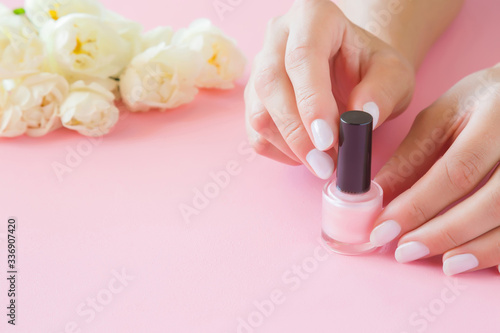 Young, perfect, groomed woman hands with nail varnish bottle. Manicure, pedicure beauty salon. Beautiful roses on pastel pink table. Fresh flowers. Close up. Empty place for text or logo. Front view.