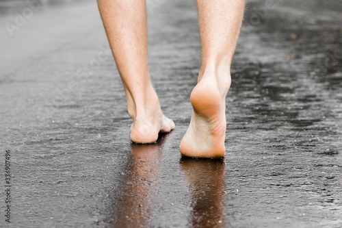 Young man barefoot walking on wet, dark black asphalt after warm rain in summer. Back view. Closeup. photo