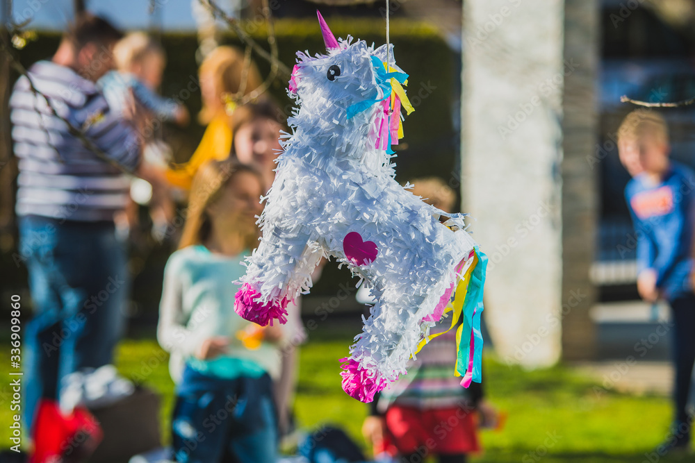 Young caucasian girl is hitting a white colored donkey shaped pinata hanging  on the tree with kids cheering in the background. Festive activity during a  birthday. Photos