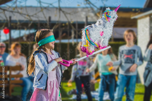 White colored donkey shaped pinata hanging on the tree with kids waiting to hit it in the background. Festive activity during a birthday