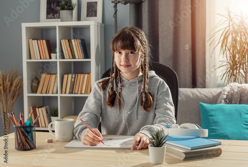 Smiling teen girl sitting at desk looking at camera, studying from home, front view photo
