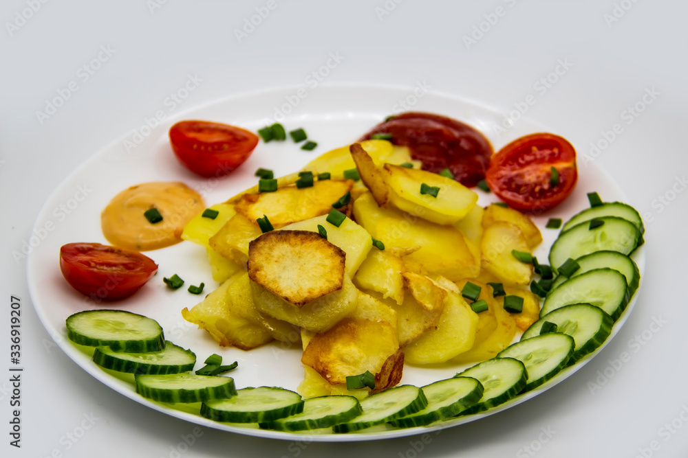 Homemade fried potatoes with fresh cucumbers, cherry tomatoes and sauces on the white plate. Delicious ukrainian fried potatoes with vegetables against background. Cooked tasty meal for lunch. Homemade fried potatoes with fresh cucumbers, cherry tomatoes and sauces on the white plate. Delicious ukrainian fried potatoes with vegetables against background. Cooked tasty meal for lunch.