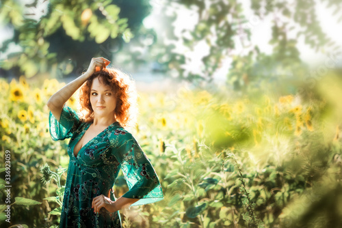Portrait of young beautiful red haired woman with one hand up in green dress in sunflowers field in back light. Summer countryside concept. Close to nature vacation. Woman and sunflowers. Summer