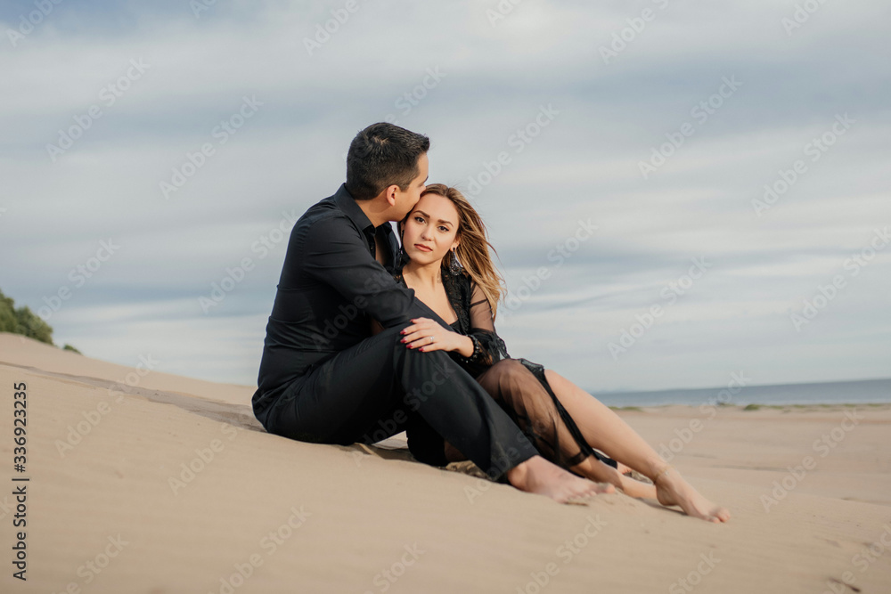 Pareja feliz sentada en la playa, relajados, casual, elegante, vestidos de negro, beso en la frente, sentados en la arena con la playa y el cielo de fondo. Abrazados. 