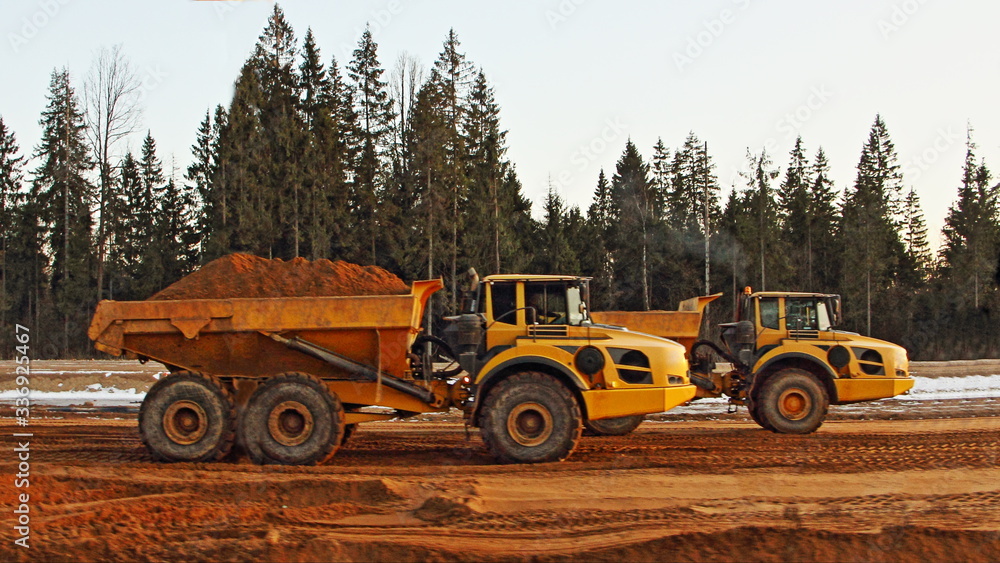 Two yellow heavy dump trucks loaded and empty drive on a construction site against the background of a trees, side view
