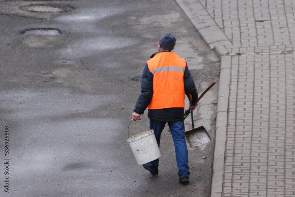 A janitor man in an orange vest walks with a bucket and scoop along an asphalt street, top view from behind A janitor man in an orange vest walks with a bucket and scoop along an asphalt street, top view from behind